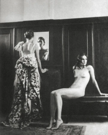 Monochrome photograph of two women indoors: one in a floral gown looking at a mirror and the other nude seated on a bench.