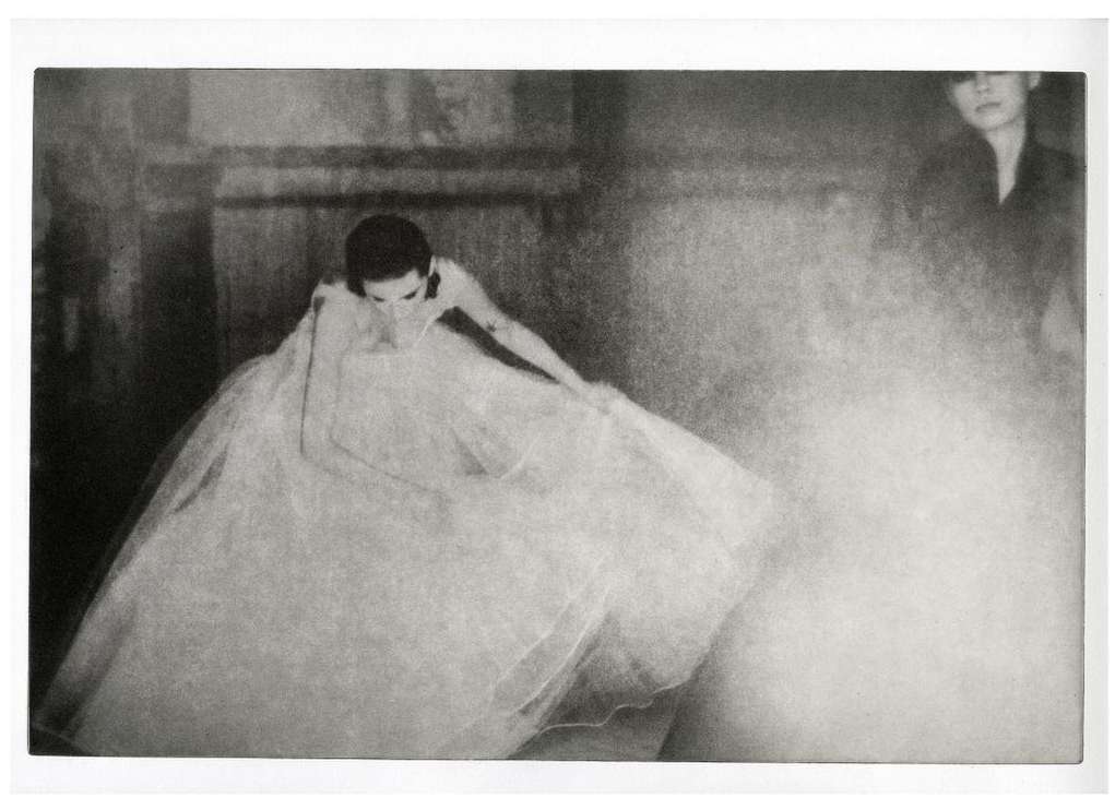 Monochrome photograph of a woman in a dress extending her arm, with another person visible in the background, creating an ethereal effect.