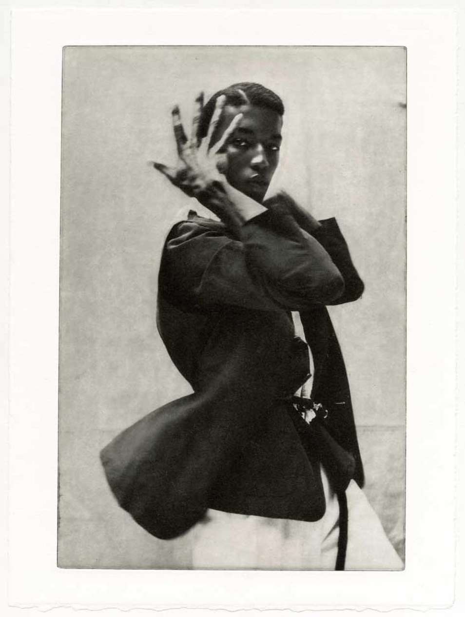 Portrait of a slender African-American man in a dark blazer and white top, posing with his hand near his face against a textured backdrop.