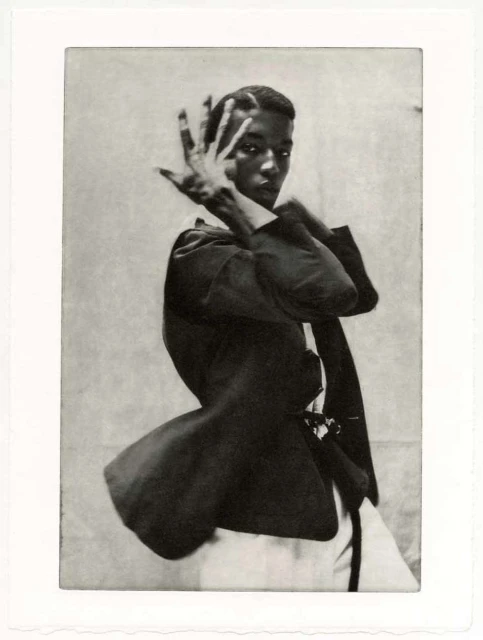 Portrait of a slender African-American man in a dark blazer and white top, posing with his hand near his face against a textured backdrop.