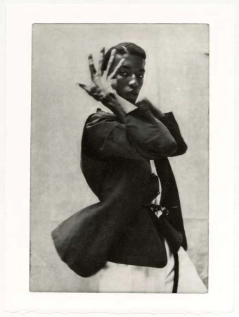 Portrait of a slender African-American man in a dark blazer and white top, posing with his hand near his face against a textured backdrop.