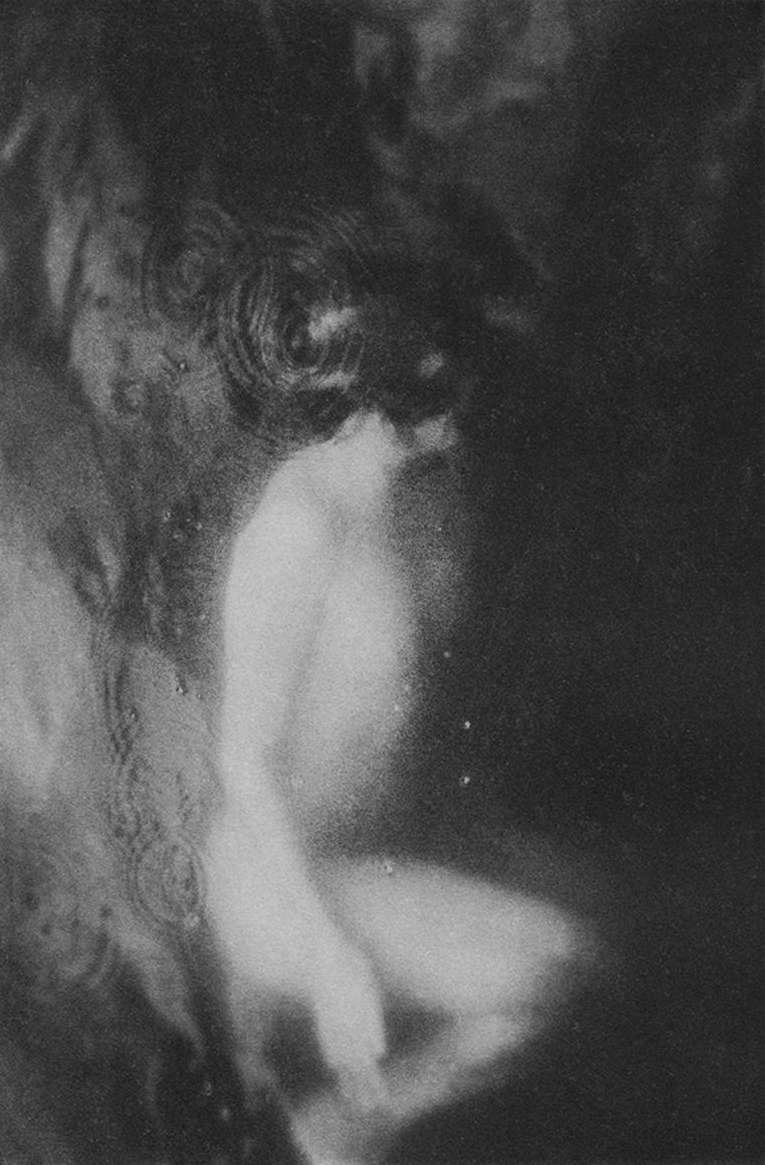 Monochrome photography of a nude female figure emerging from a dark, blurred background, suggestive of a dreamlike or ethereal quality.