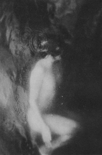 Monochrome photography of a nude female figure emerging from a dark, blurred background, suggestive of a dreamlike or ethereal quality.