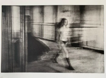 Monochrome photographic image of a woman walking through an interior space captured with long exposure motion blur