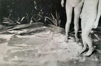 Monochrome photograph of two nude figures standing near painted backdrop with foliage and textured ground by Chantal Elisabeth Ariens