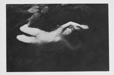 Monochrome underwater photograph of a figure swimming beneath lily pads in dark water by Chantal Elisabeth Ariens
