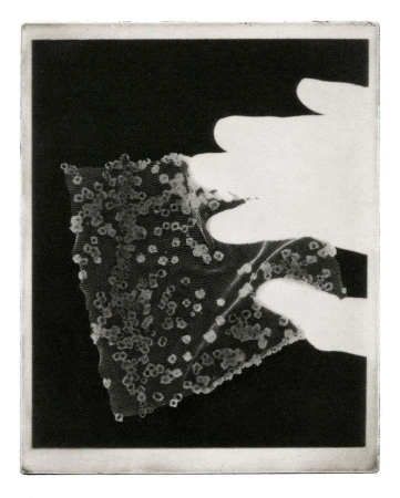 Monochrome photographic print showing hands holding a textured textile fragment covered in small particulate crystals.