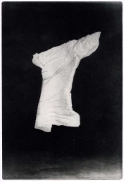 Monochrome photographic print of a draped white fabric sculpture suspended against a dark textured background