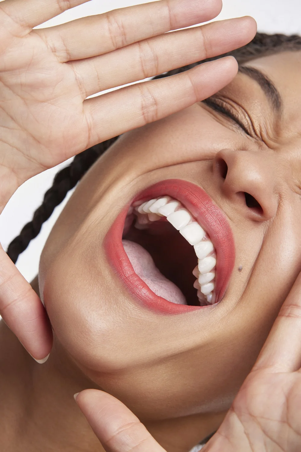 Close-up studio shot of a laughing woman with braided hair, pink lipstick, and hands framing her face against a white backdrop.