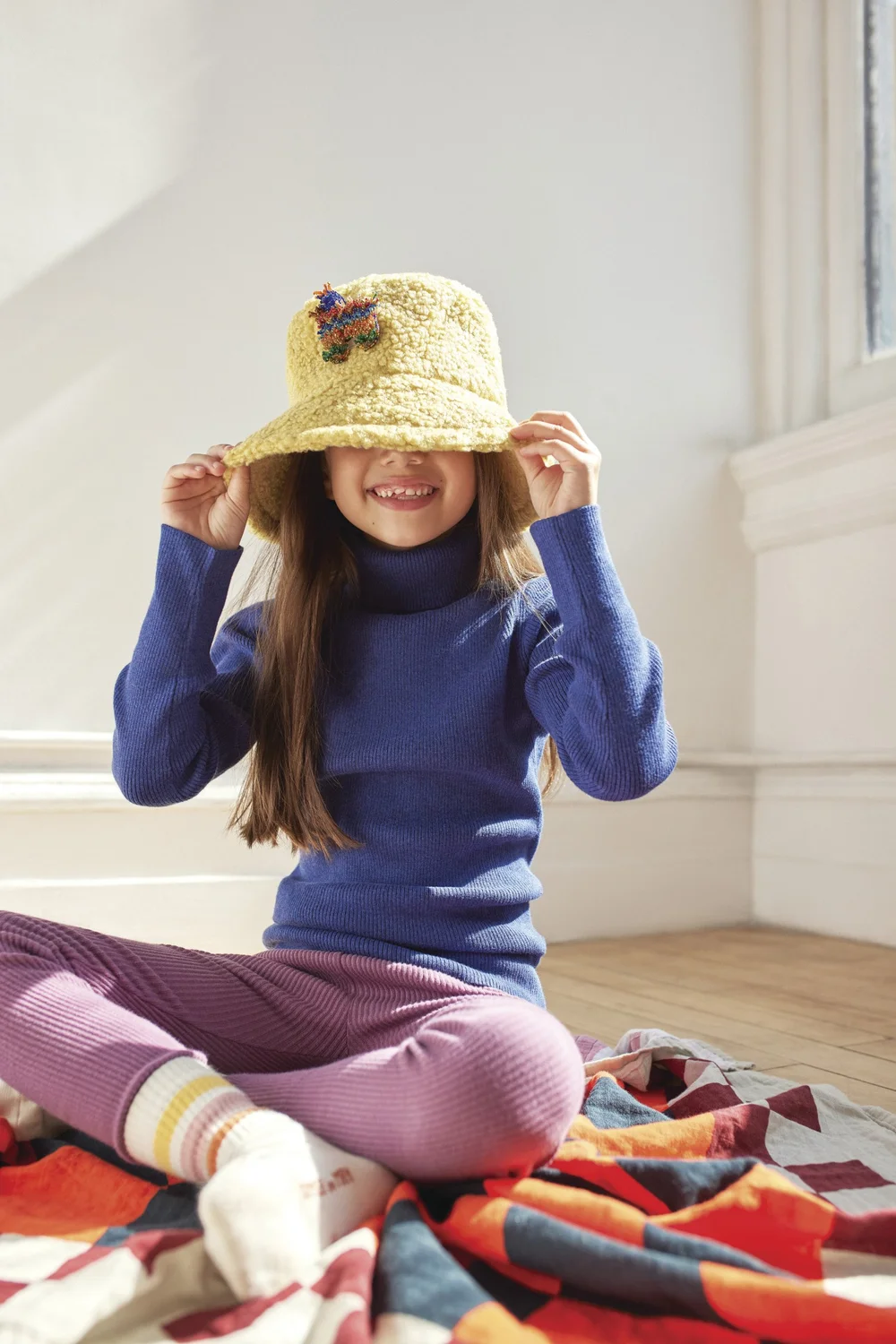 Smiling girl in blue turtleneck hides face with yellow crochet bucket hat, sitting on patterned blanket in a sunlit room.