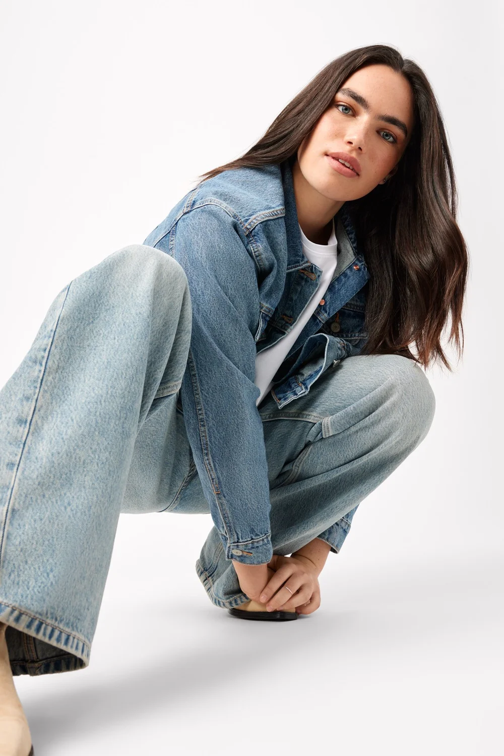 Woman crouches in denim jacket and jeans, smiling in a bright photo studio against a white backdrop; fashion editorial photography.