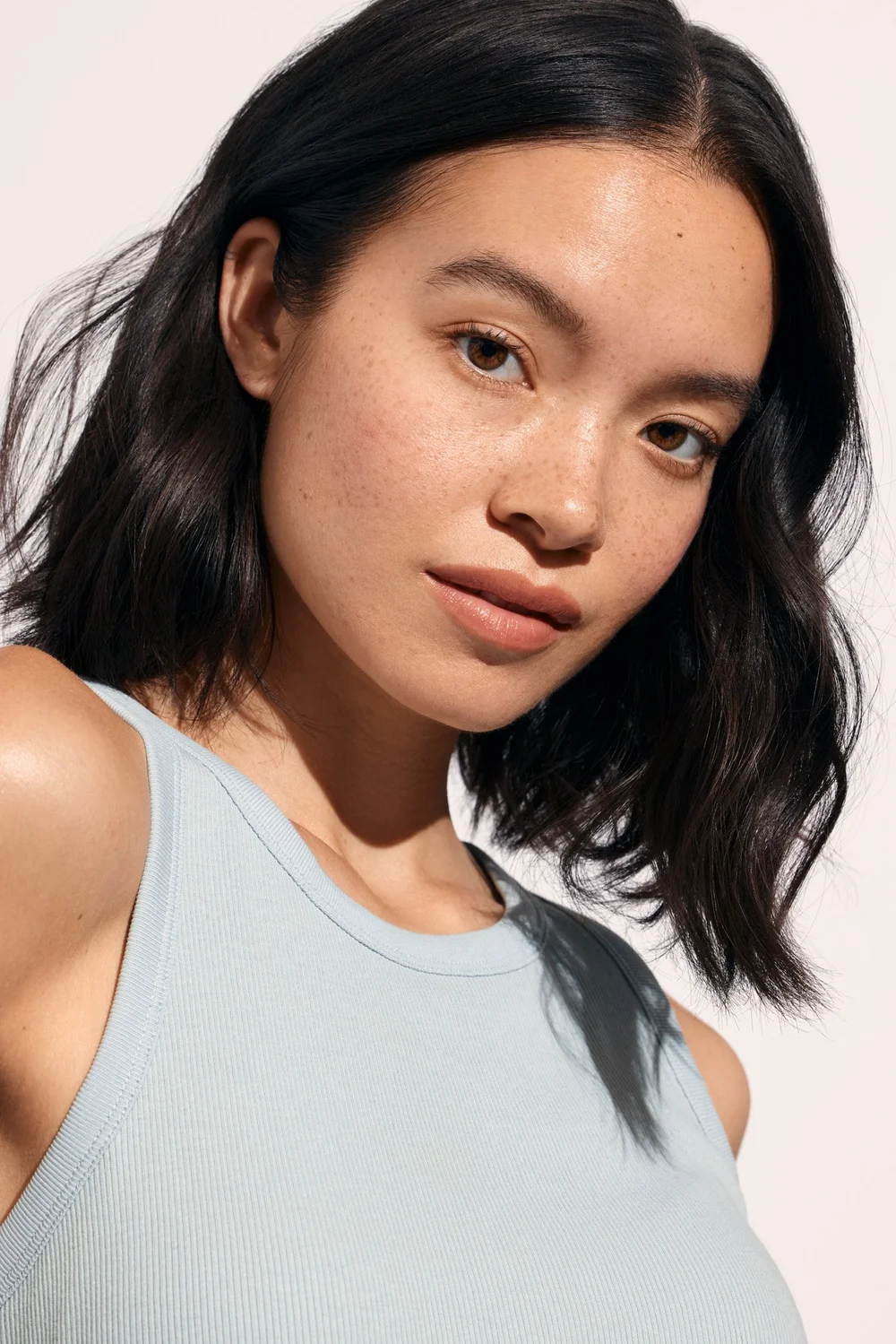 Close-up portrait of a young adult woman with short wavy dark hair, brown eyes, light skin, wearing a light blue ribbed tank top.