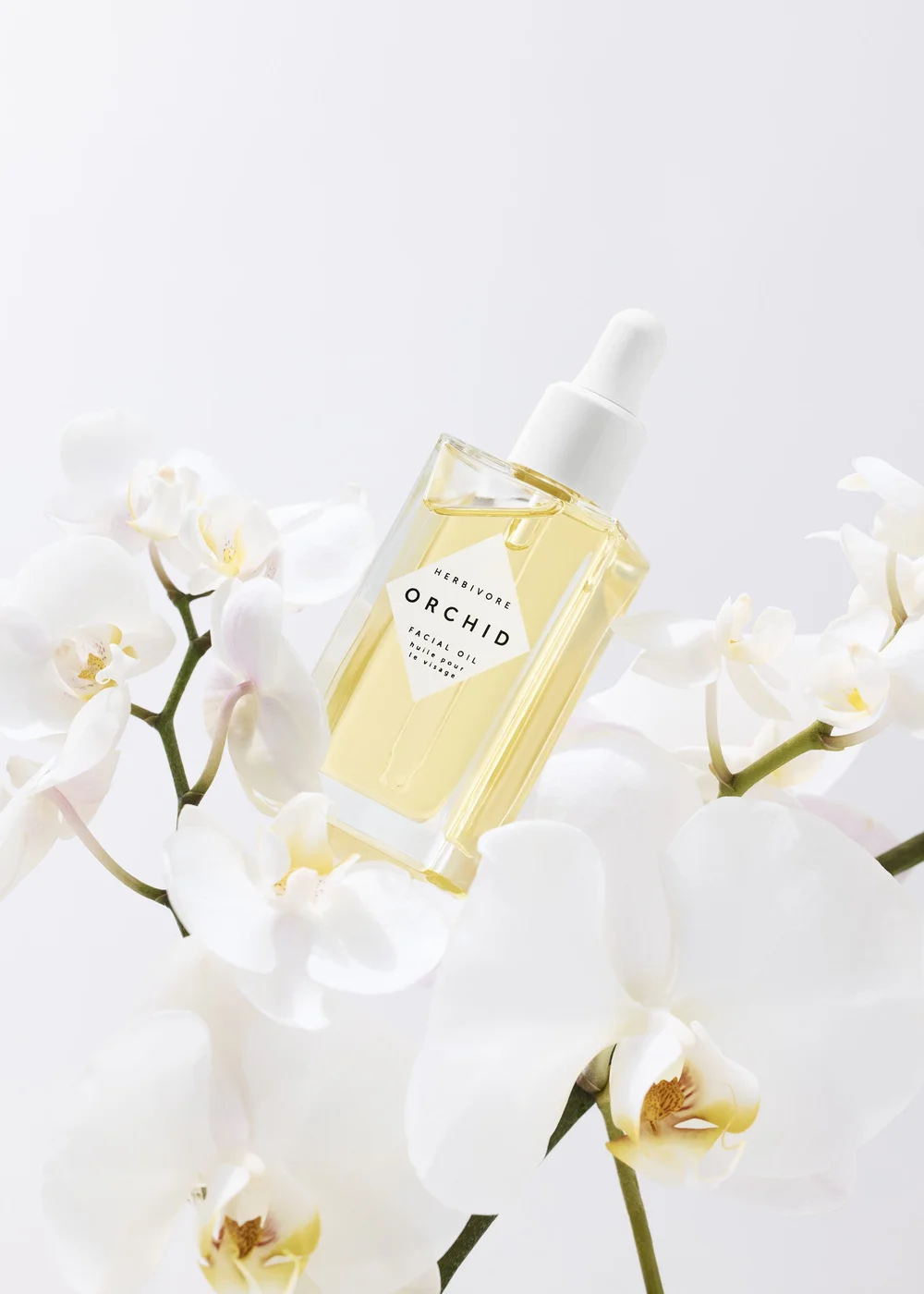 Square bottle of Herbivore Orchid Facial Oil with dropper top surrounded by delicate white orchid blossoms on a white background.