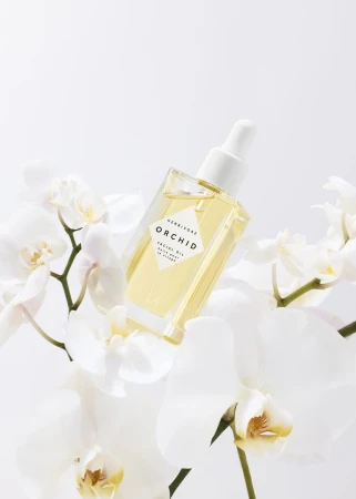 Square bottle of Herbivore Orchid Facial Oil with dropper top surrounded by delicate white orchid blossoms on a white background.