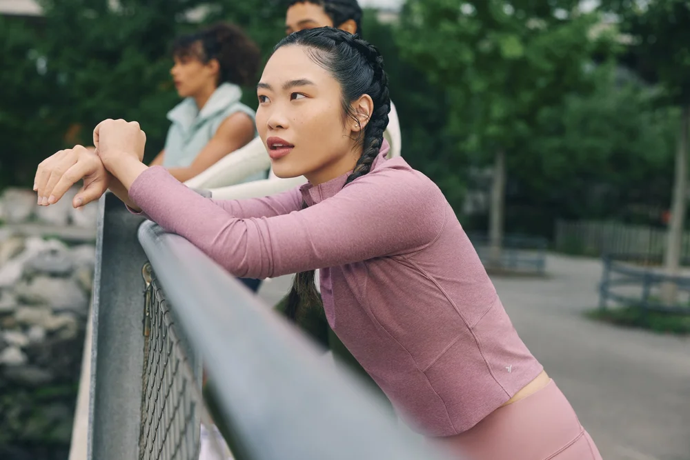 Young woman in athleisure wear leans on railing in urban park setting with two other women in background, daytime, medium shot.