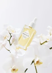 A glass bottle of yellow facial oil nestled among delicate white orchid flowers against a plain white background.