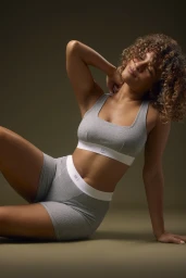 Woman with curly hair wearing grey athletic bra and shorts sitting against dark background