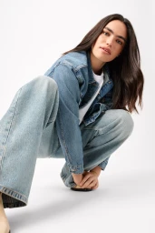 Woman crouching wearing a blue denim jacket and baggy jeans against a white seamless background