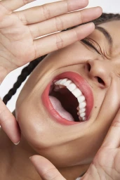 Close-up portrait of a woman with braided hair shouting with her mouth wide open, framing her face with both hands against a white background.