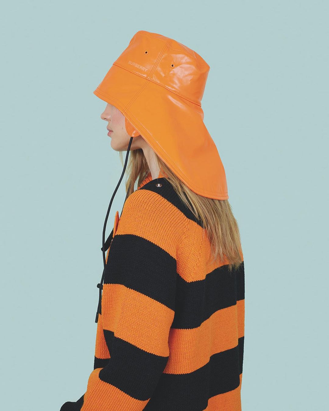 A woman models a vinyl orange rain hat with ties and a striped orange and black sweater on a light blue background in a studio setting.