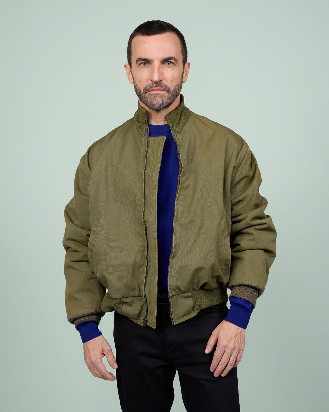 Adult male in an olive green jacket poses indoors against a light green wall. He is wearing a blue top and dark trousers.