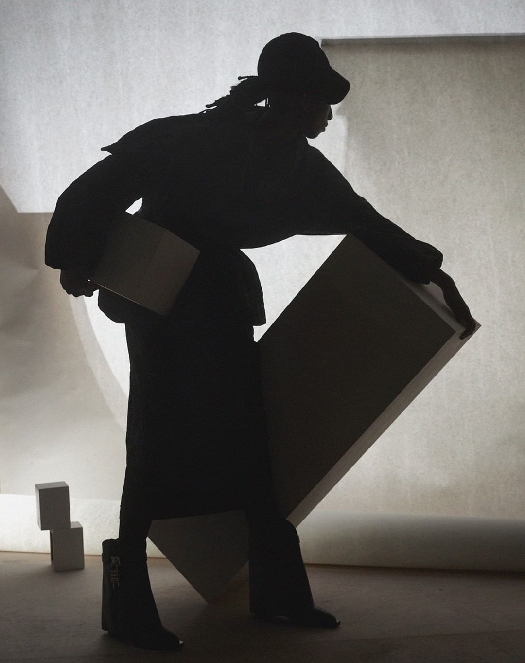 Silhouette of a person in streetwear, wearing a cap and boots, carrying a flat object against a light backdrop with geometric shapes.