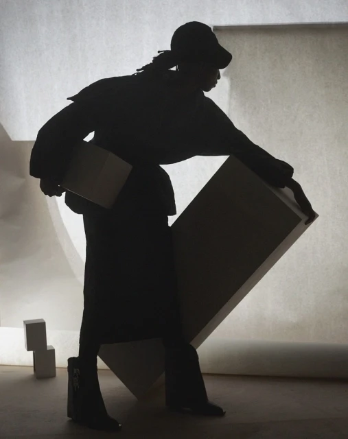 Silhouette of a person in streetwear, wearing a cap and boots, carrying a flat object against a light backdrop with geometric shapes.