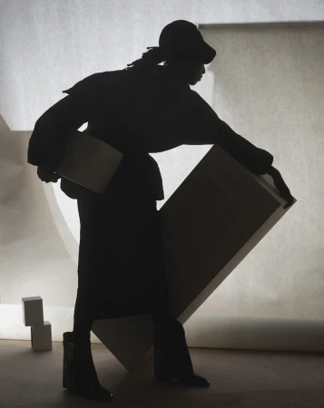 Silhouette of a person in streetwear, wearing a cap and boots, carrying a flat object against a light backdrop with geometric shapes.