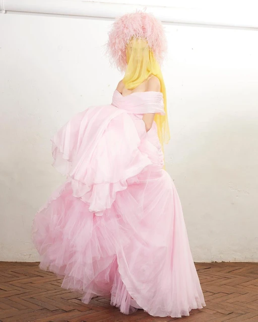 Woman in an elaborate pink tulle gown and feathered headpiece stands against a white wall, creating a dreamy, high-fashion editorial image.