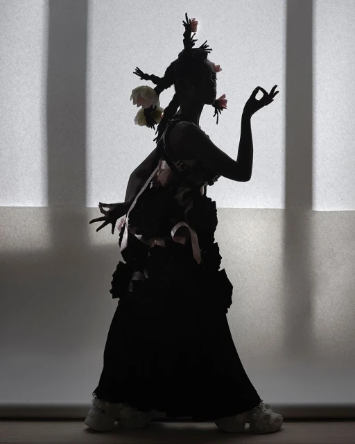 Silhouette of a model in an avant-garde outfit, adorned with floral elements, striking a pose against a softly lit white backdrop.