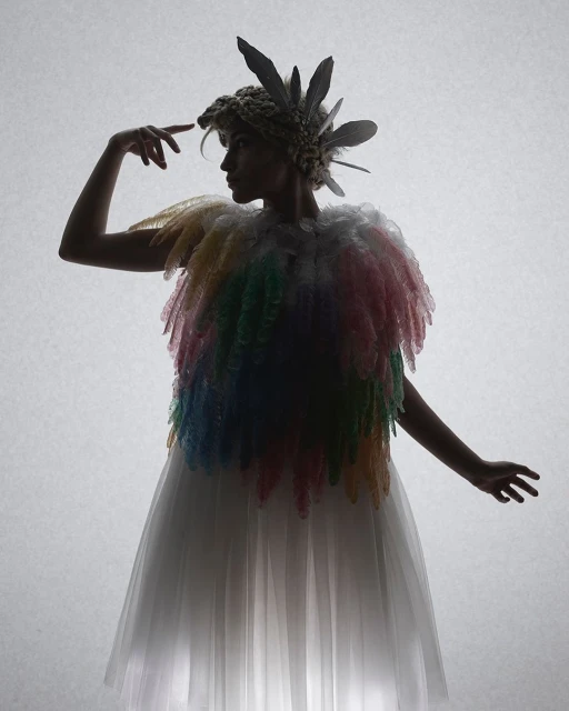 Person in feathered couture gown and headdress stands in a photo studio with backlight. Fashion, beauty and editorial photography.