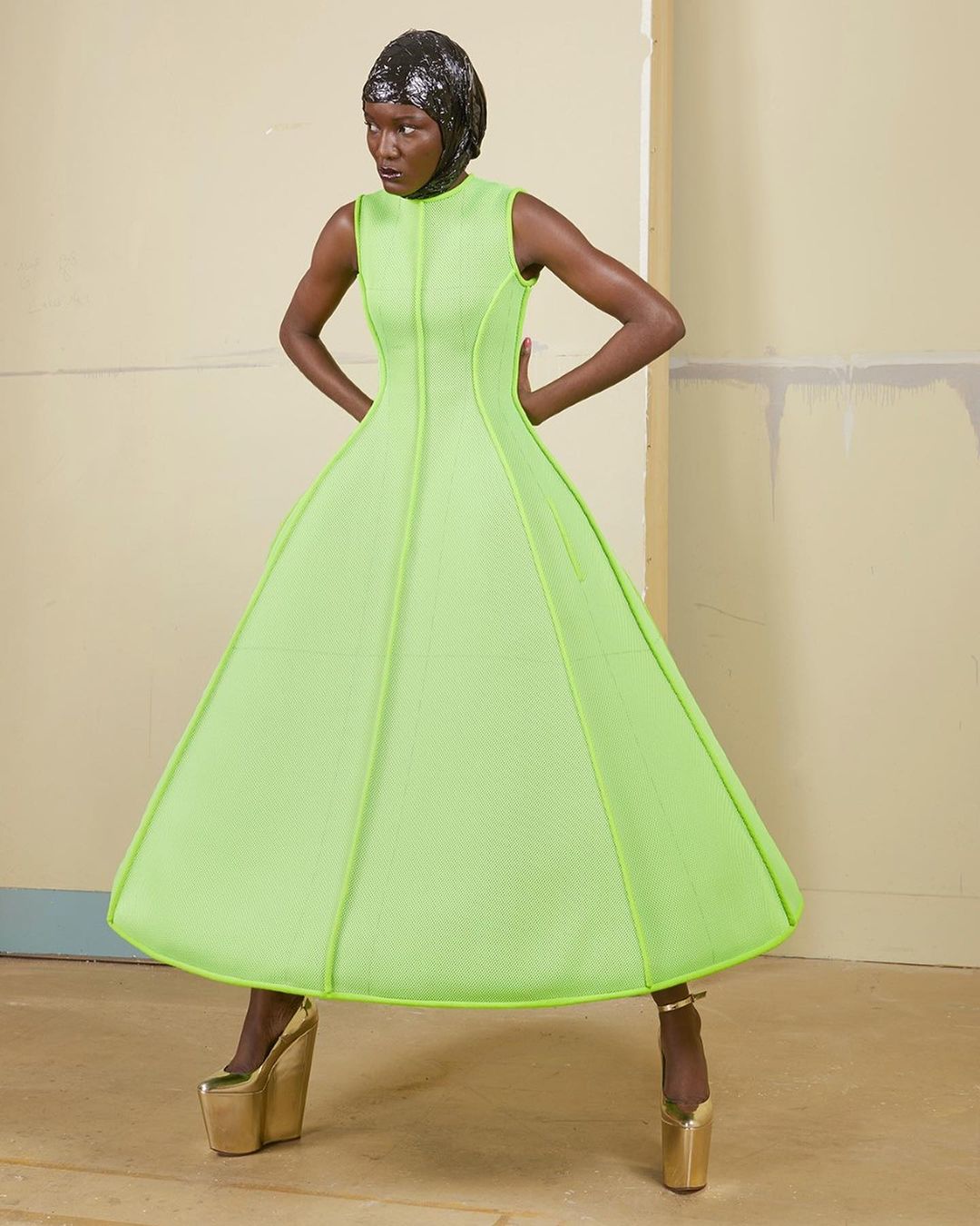 Fashion shot of a Black woman wearing a neon green dress with a matching head wrap and gold platform heels posing in a studio.