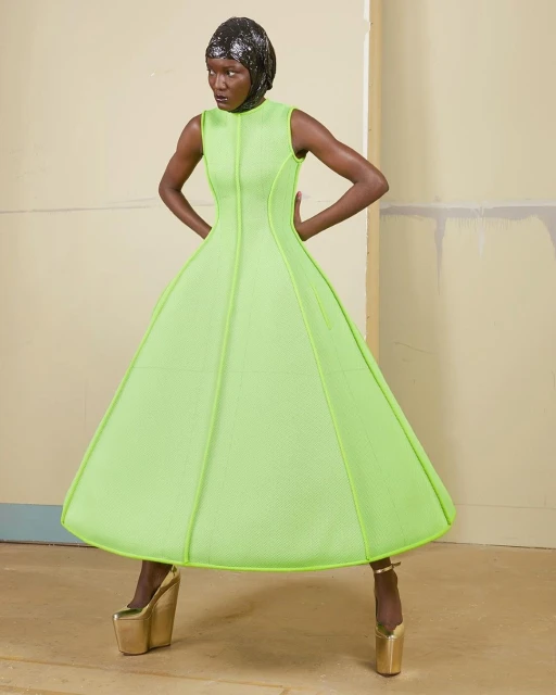 Fashion shot of a Black woman wearing a neon green dress with a matching head wrap and gold platform heels posing in a studio.