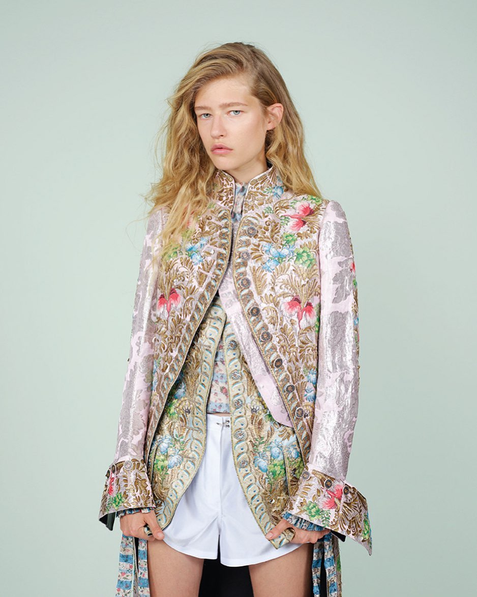 A fair-skinned woman with long blonde hair poses in an embroidered pink jacket and white shorts against a light green background.