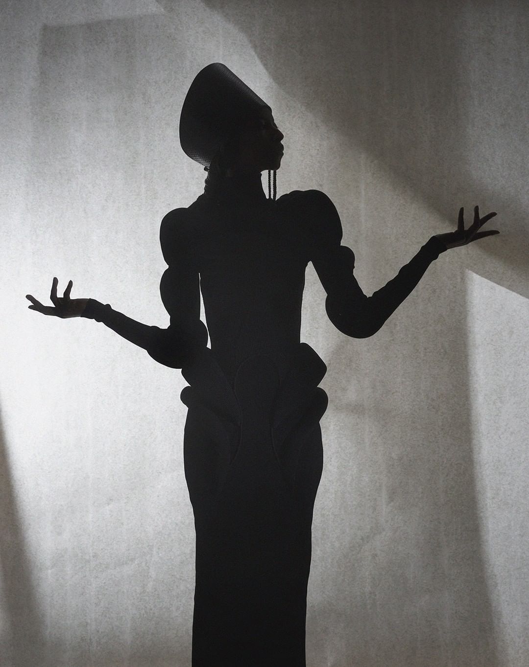 Silhouette of a woman in a vintage dress and headdress stands with her arms outstretched against a textured studio background.
