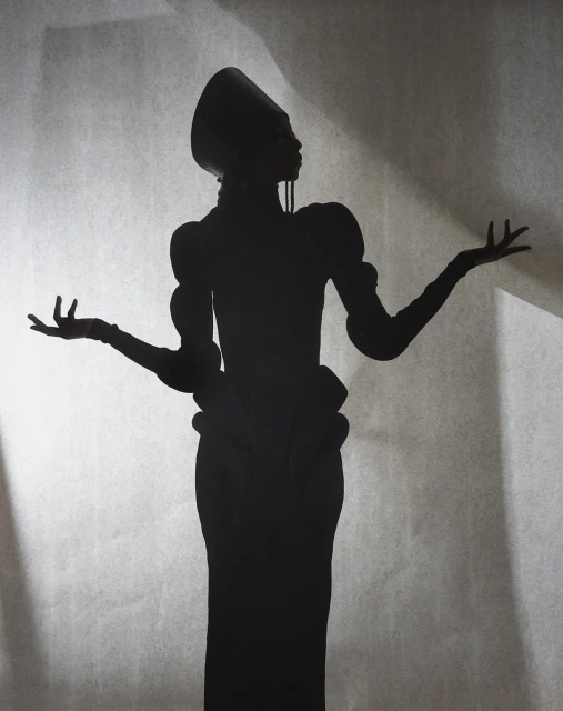Silhouette of a woman in a vintage dress and headdress stands with her arms outstretched against a textured studio background.