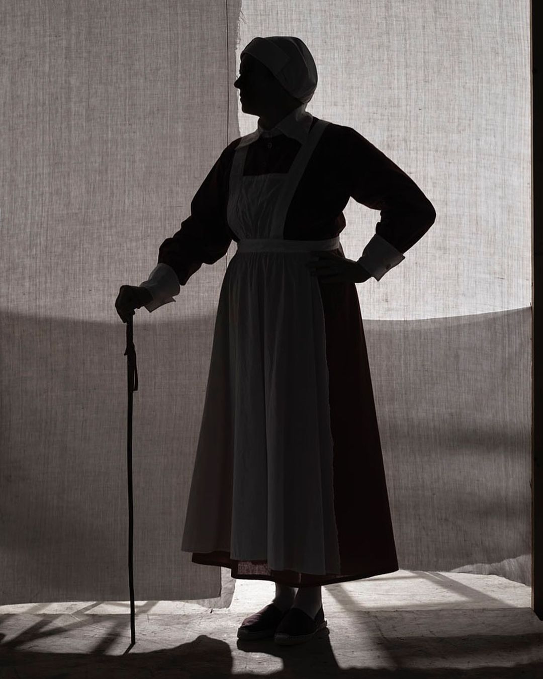 Silhouette of an adult woman in vintage clothing holding a cane, standing in front of a textured gray backdrop with light shining through.