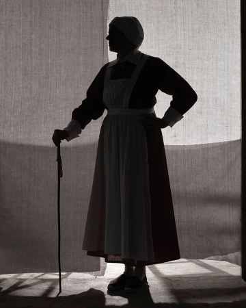 Silhouette of an adult woman in vintage clothing holding a cane, standing in front of a textured gray backdrop with light shining through.