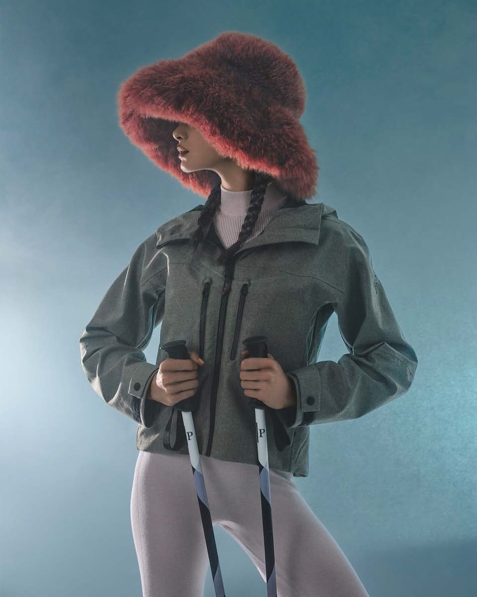 Studio shot of an adult female wearing a gray jacket, fur hat, and holding ski poles against a blue gradient background, posing fashionably.