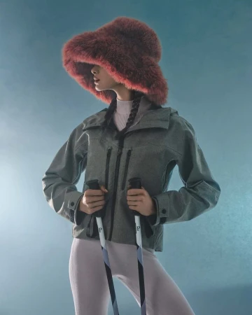 Studio shot of an adult female wearing a gray jacket, fur hat, and holding ski poles against a blue gradient background, posing fashionably.