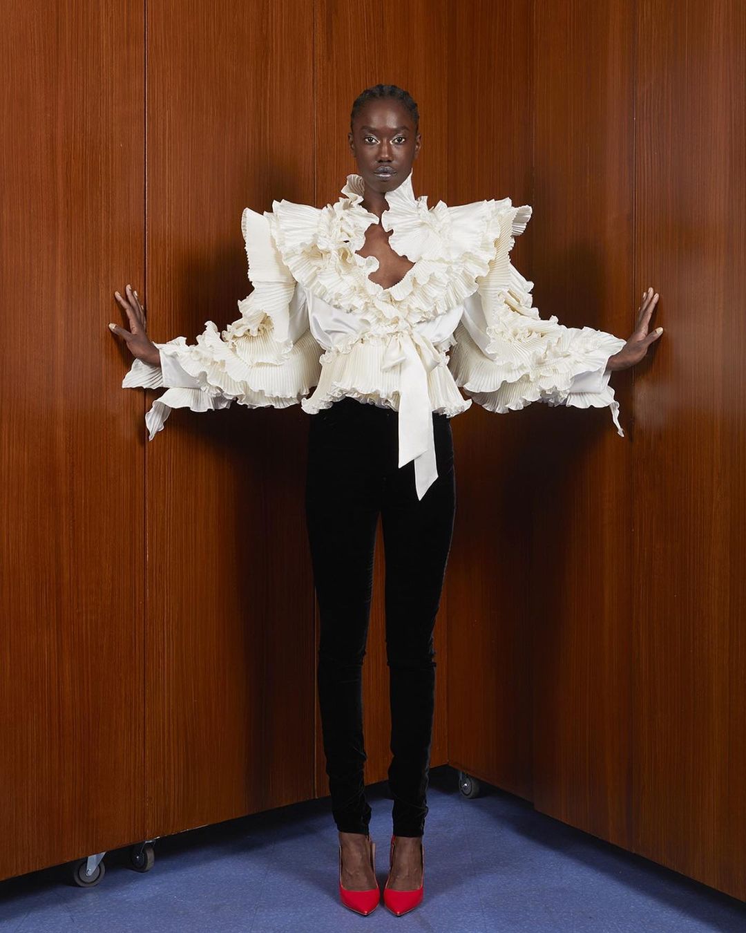 Black woman in a white ruffled blouse and dark trousers standing against wood paneling by Charles Freger