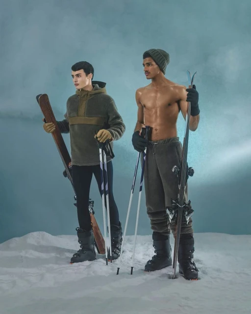 Two athletic men pose in winter ski outfits against a blue backdrop, holding skis and poles, embodying a blend of fitness and fashion.