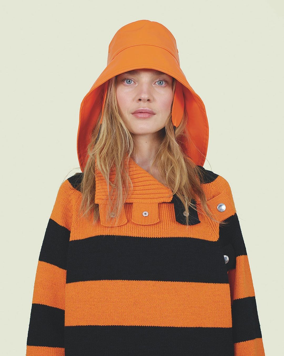 A fair-skinned woman with blonde hair poses indoors, wearing an orange sunhat and a bold orange and black striped sweater.
