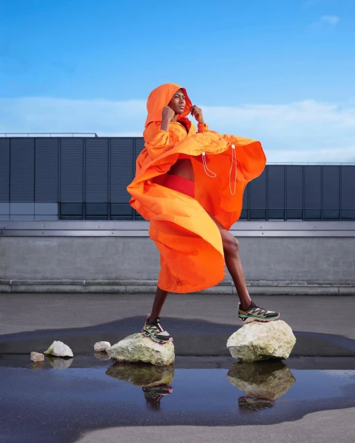 A person in an orange hooded garment stands on rocks near water with a modern building and blue sky in the background on a sunny day.