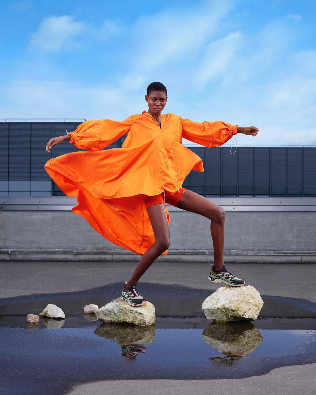 An adult Black female fashion model in an orange outfit balances on rocks near a puddle in an outdoor urban environment under a blue sky.