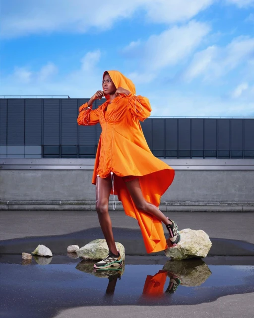 An adult woman in an orange dress and sneakers stands on rocks near a puddle outdoors, with a blue sky and building in the background.