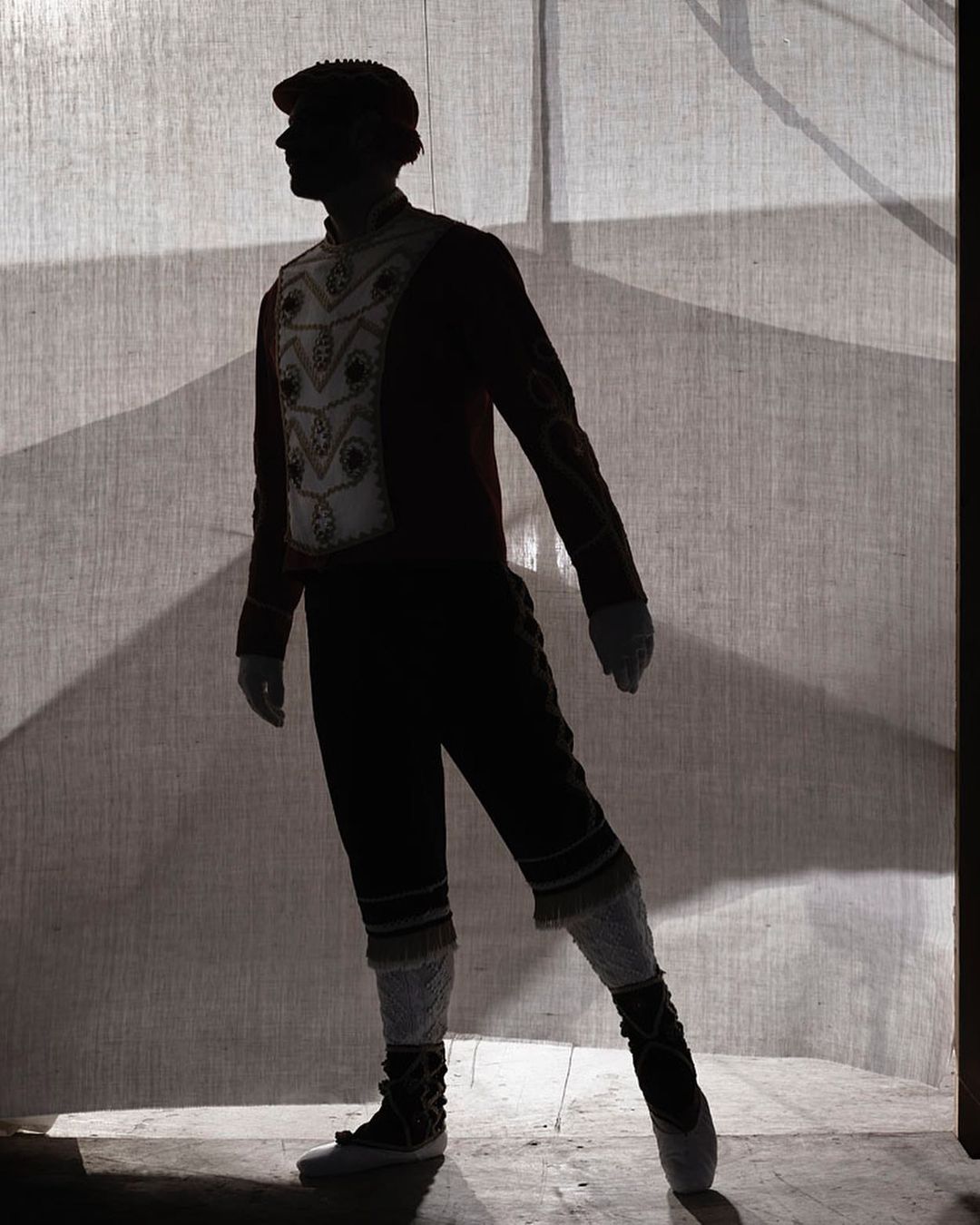 A silhouette of a single ballet dancer in an ornate costume on a stage standing in front of a textile backdrop.