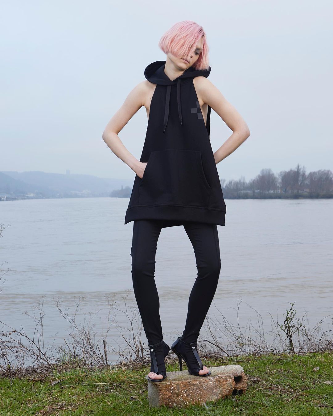 A woman with pink hair poses in black sleeveless hoodie, leggings, and high-heeled sandals outdoors near a river under overcast skies.