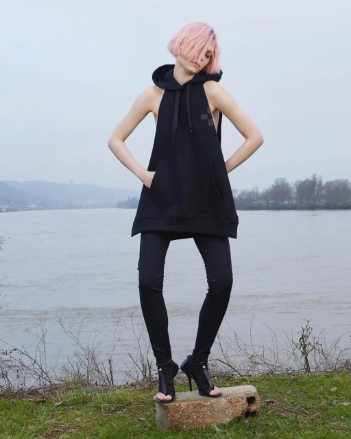 A woman with pink hair poses in black sleeveless hoodie, leggings, and high-heeled sandals outdoors near a river under overcast skies.