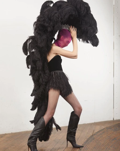 Model in a black strapless dress, feathered skirt, and boots supports a large black feathered headdress and pink face covering in a studio.